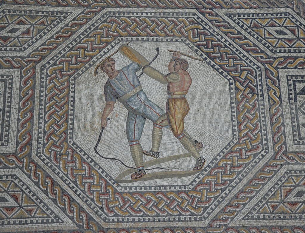 Octagonal medallion depicting two combatants attacking one another with cudgels and a whip. The introduction to the gladiatorial contests consisted of a prolusio (prelude). The various pairs fought with blunted weapons, giving the foretaste of their skills.